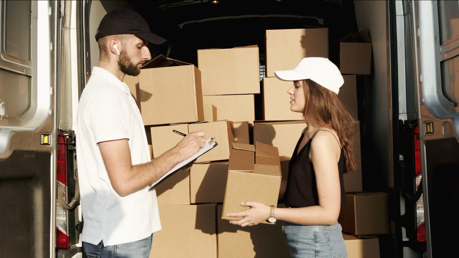A white delivery van with its rear doors open, loaded with parcels, as a courier checks a consignment off